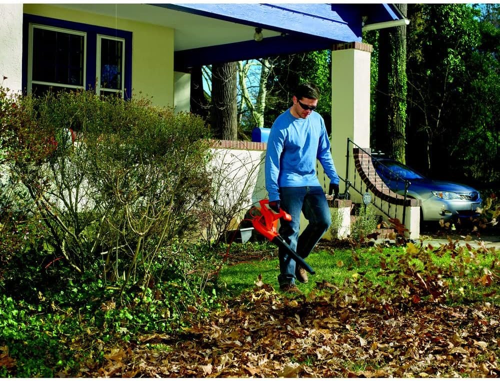 A man in a blue shirt and jeans using the BLACK+DECKER electric leaf blower to clear autumn leaves from a grassy front yard.